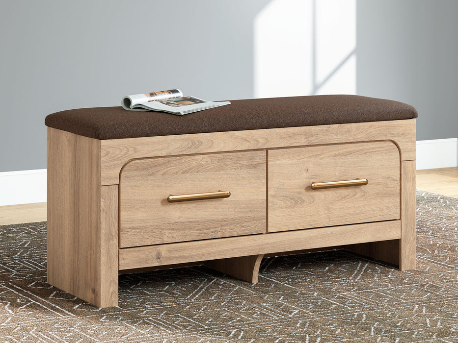Tan upholstered storage bench by Signature Design Ashley, featuring a cushioned top and wooden legs in a cozy bedroom setting.